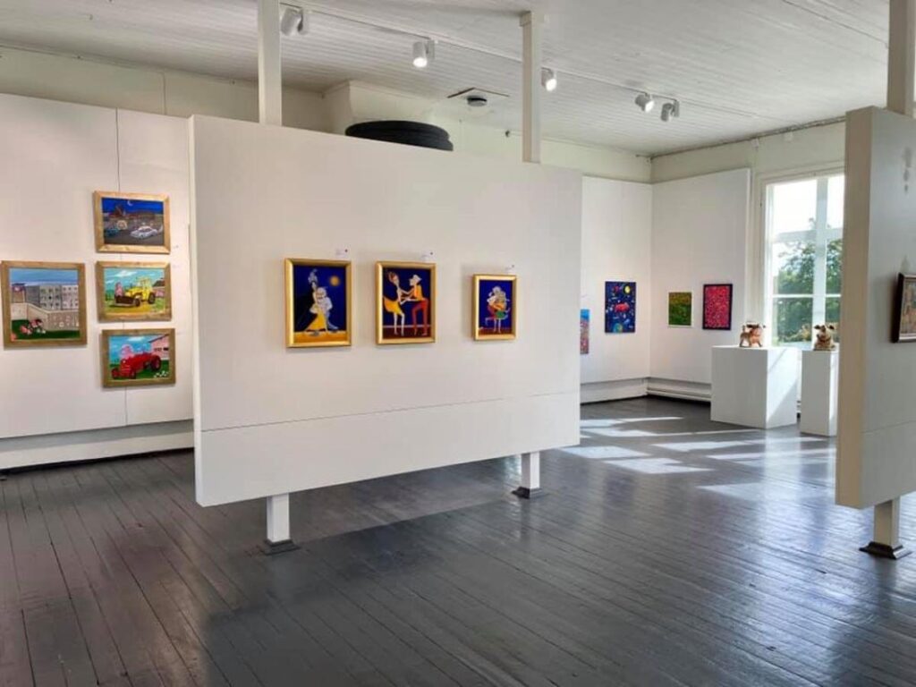 Una sala de galería luminosa. Exhibiendo arte naíf colorido con pinturas alegres de escenas al aire libre, figuras danzantes y esculturas de perros en pedestales.